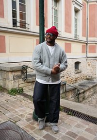 Hombre con gorro rojo, cárdigan gris, camiseta blanca y vaqueros oscuros apoyado en un farol verde en una calle empedrada.