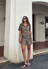 Macacão de manga curta com padrão de leopardo em tons de bege e castanho, com colarinho e dois bolsos frontais, combinado com sandálias pretas.
