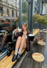 Une personne assise dans un manteau marron tient une tasse à thé blanche, portant des chaussettes grises et des mocassins noirs, avec des bancs en bois et des petites tables à proximité.