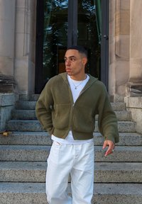 Cárdigan de canalé con cremallera en verde oliva, con bolsillos delanteros, llevado sobre una camiseta blanca, combinado con pantalones blancos holgados, de pie sobre escalones de piedra.