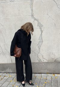 Persoon in een zwarte jas en wijde broek, houdend een bruine handtas, staand op een met bladeren bezaaid trottoir tegen een gebroken beige stenen muur.