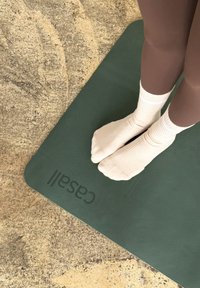 Zelená jógová podložka s texturovaným povrchem, na které je zobrazená značka "Casall". Bílé ponožky na nohou položených na podložce.