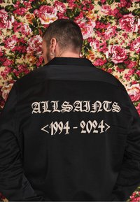 Camisa preta com o texto "ALLSAINTS <1994 - 2024>" em branco, exibido de forma proeminente nas costas. Textura de tecido suave.