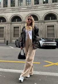 Μοντέλο φοράει ένα μαύρο δερμάτινο oversized τζάκετ, λευκή cropped μπλούζα, μπεζ παντελόνι με φαρδιές λεπτομέρειες και ασημένια αθλητικά παπούτσια, κρατώντας μια μικρή μαύρη τσάντα.
