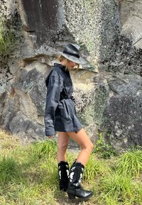 Giubbotto in denim nero con vita sfiancata, abbinato a stivali da cowboy neri con accenti floreali bianchi. Ambientato su uno sfondo roccioso.