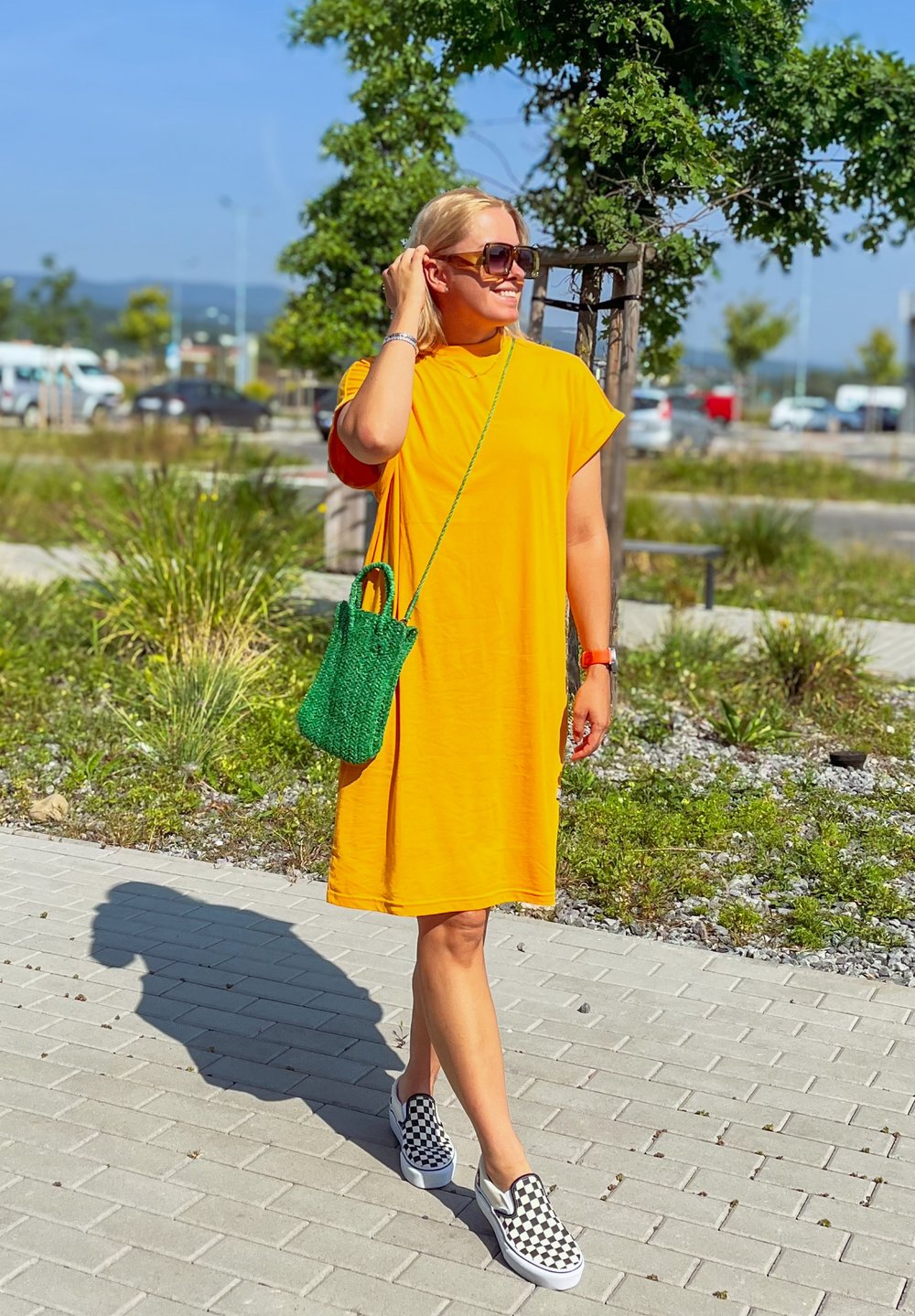 Vestido amarelo de comprimento médio com mangas curtas, acessorizado com uma mala de vime verde e sapatos slip-on às manchas. Cenário ao ar livre ensolarado.