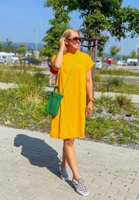 Vestido amarelo de comprimento médio com mangas curtas, acessorizado com uma mala de vime verde e sapatos slip-on às manchas. Cenário ao ar livre ensolarado.