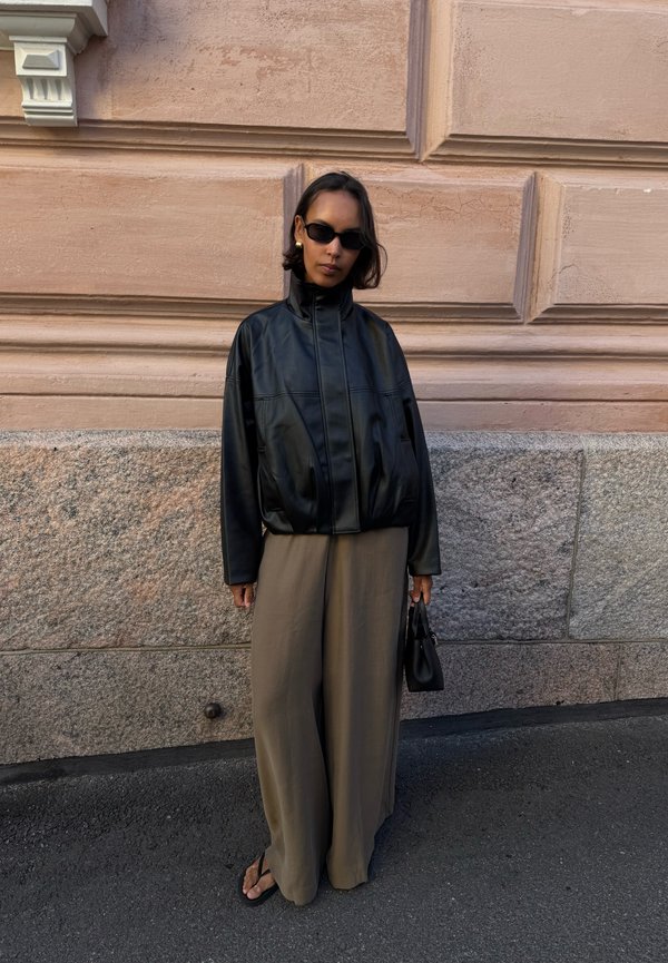 Persona que lleva una chaqueta de cuero negra, gafas de sol oscuras, pantalones beige holgados y sandalias, de pie contra una pared texturizada de piedra y color melocotón, sosteniendo un bolso negro.