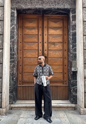 Camisa de manga corta con estampado en blanco y negro, pantalones negros sueltos, sosteniendo un vaso aislante blanco frente a unas puertas dobles de madera.