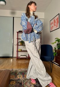 Μπλε σακάκι με λουλουδάτο σχέδιο και λευκές λεπτομέρειες, συνδυασμένο με λαμπερά ασημένια παντελόνια και ροζ αθλητικά παπούτσια. Το φόντο περιλαμβάνει φυτά και έργα τέχνης.