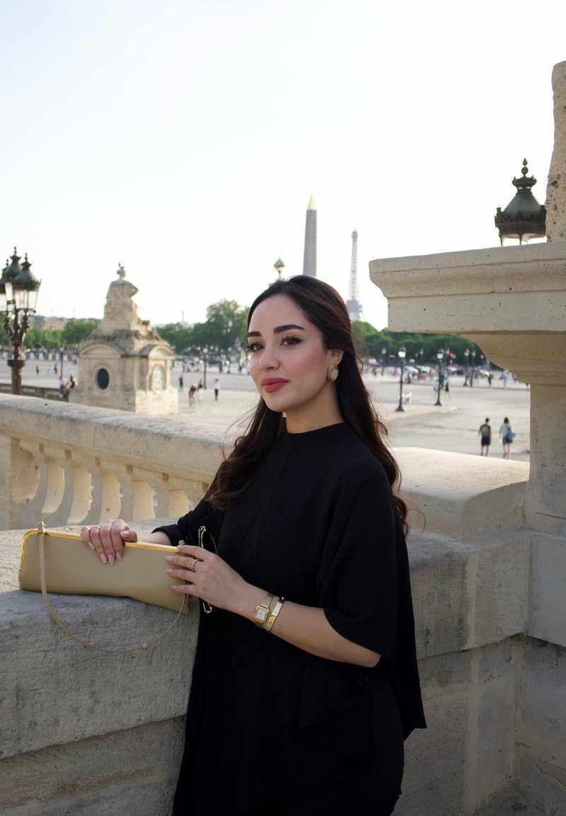 Femme en robe noire tenant une pochette beige, debout sur un balcon en pierre avec la tour Eiffel et un obélisque en arrière-plan par une journée ensoleillée.