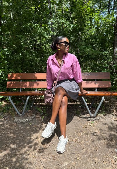 Un banco de madera marrón con patas de metal en un entorno forestal verde. La persona lleva una camisa a rayas rosa, una falda gris y zapatillas blancas.
