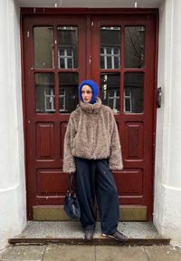 Veste surdimensionnée en fourrure marron avec col montant, associée à un pantalon noir ample et un chapeau bleu tricoté. Sac noir et chaussures marron complètent le look.