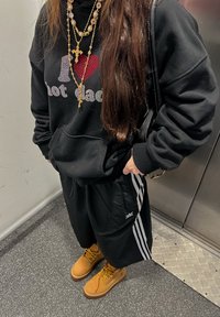 Čierna mikina s nápisom "I ❤️ hot dads", v kombinácii s čiernymi šortkami s bielymi pruhmi. Béžové čižmy a vrstvené zlaté náhrdelníky dotvárajú outfit.