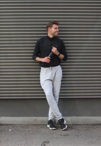 Abbigliamento sportivo: maglietta nera a maniche lunghe, pantaloni della tuta grigi con tasche e sneakers nere. In piedi contro un muro di metallo ondulato, con una bottiglia d'acqua in mano.