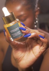 Przezroczysta butelka lśniącego złotego olejku do włosów i ciała z białą pipetą. Etykieta zawiera tekst w białym i zielonym kolorze, trzymana w ręce z długimi niebieskimi paznokciami.