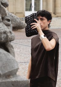 Persoon met kort krullend haar houdt een zwarte geweven clutch dicht bij het gezicht, draagt een donker geplooide top en een zilveren armband, staand bij een stenen beeldhouwwerk.