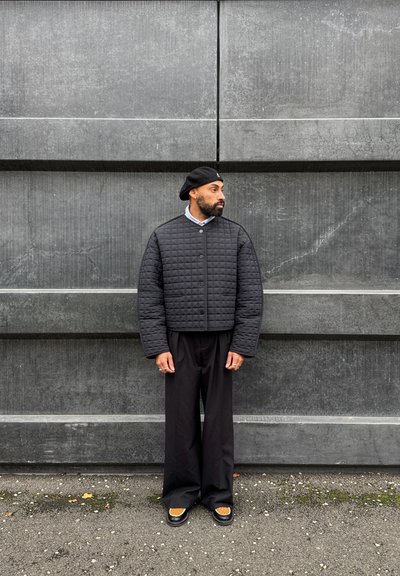 Chaqueta acolchada negra con cuello redondo, sobre una camisa de color azul claro, combinada con pantalones sueltos negros y zapatos con detalles marrones.
