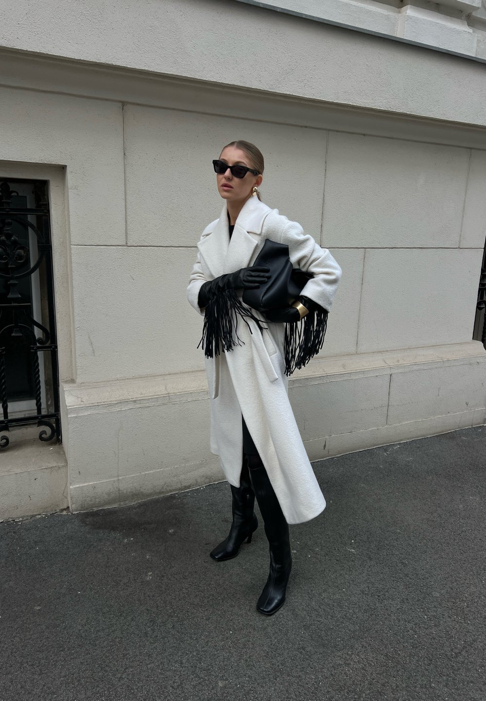 Λευκό υπερμεγέθες παλτό από υφασμάτινη υφή, μαύρα δερμάτινα γάντια με κρόσσια, μαύρη τσάντα, κομψά μαύρα μποτάκια και σκούρα γυαλιά ηλίου.