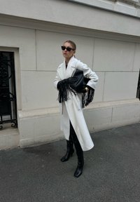 Λευκό υπερμεγέθες παλτό από υφασμάτινη υφή, μαύρα δερμάτινα γάντια με κρόσσια, μαύρη τσάντα, κομψά μαύρα μποτάκια και σκούρα γυαλιά ηλίου.