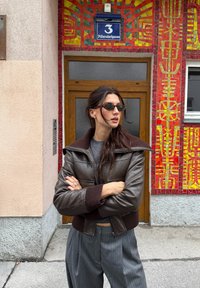 Hnědá kožená bomber bunda se širokým límcem a žebrovanými manžetami, nošená s šedými kalhotami s pruhy. Mosaikový vzor na pozadí.
