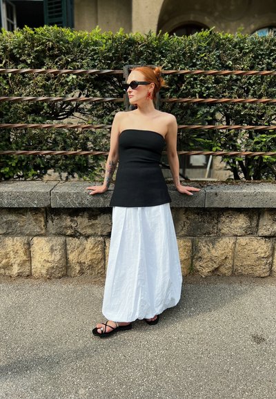 Top sin tirantes negro, falda larga fluida blanca, sandalias planas negras, pendientes rojos, de pie contra una pared de piedra con un fondo de vegetación.