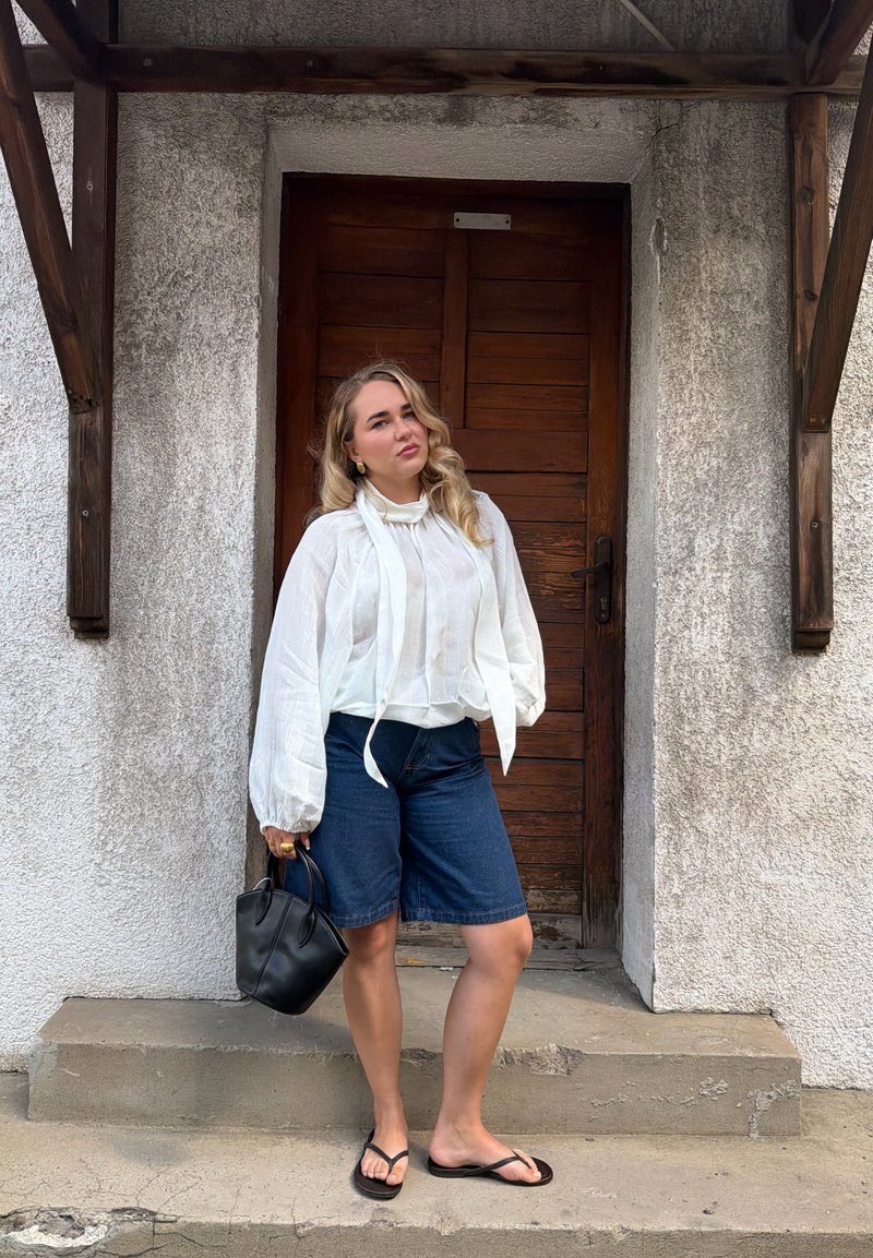 Femme aux longs cheveux blonds portant un chemisier blanc, un short bleu, des sandales noires et tenant un sac à main noir, debout sur des marches devant une porte en bois.