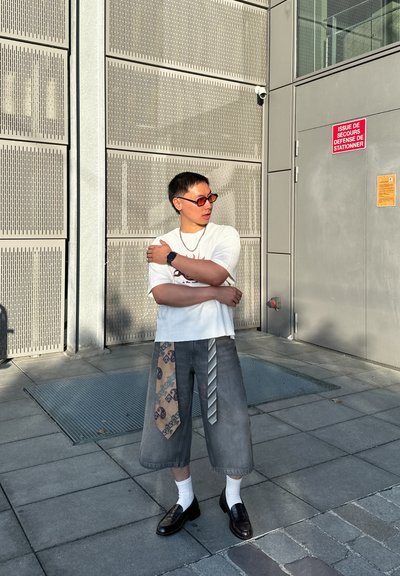 Camiseta blanca corta, pantalones cortos grises de pierna ancha con un acento estampado, mocasines negros, calcetines blancos, gafas de sol y un reloj. Fondo texturizado.