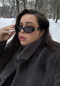 Γυναίκα με μακριά σκούρα μαλλιά που φοράει μαύρα γυαλιά ηλίου και μαύρο παλτό από γούνα σε χιονισμένο πάρκο με γυμνά δέντρα στο βάθος.