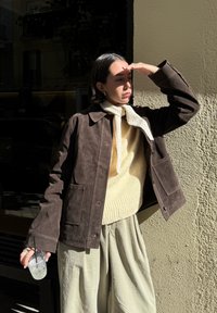 Brown suede jacket with a collar, featuring two front pockets, layered over a cream knitted sweater. The model is holding a clear cup with a straw and is dressed in loose-fitting light-coloured trousers.