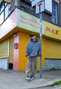 Joven con sudadera gris y pantalones beige apoyado en un poste frente a la cerrada tienda East Wall Pizza Max de color amarillo.