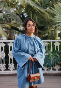 Robe en denim avec de longues manches larges et un col froncé ; associée à un sac bandoulière marron avec un logo. Entourée de verdure.