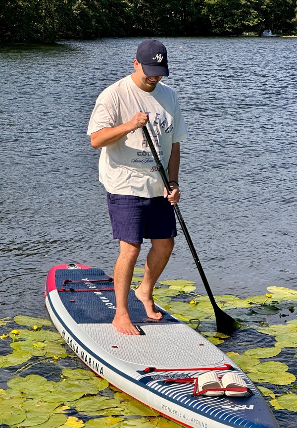 Un homme pieds nus fait du paddle à travers des nénuphars sur un lac bordé d’arbres.