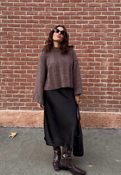 Jersey marrón corto, vestido midi de satén negro, botas marrones de piel sintética hasta la rodilla y bolso negro contra un fondo de pared de ladrillo.