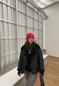 Μαύρο φουσκωτό μπουφάν με συνθετική γούνα στον γιακά, γκρι παντελόνι και λαμπερή κόκκινη σκούφια. Η φιγούρα κρατά μια μαύρη τσάντα σε έναν μοντέρνο εσωτερικό χώρο.