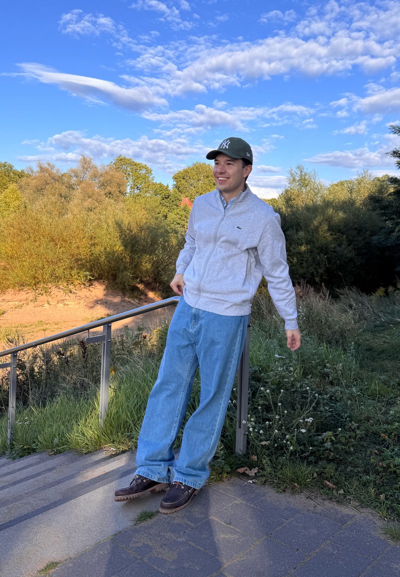 Jovem vestido com uma jaqueta cinza com fecho, calças de ganga azuis e um chapéu escuro, encostado a um corrimão perto de escadas exteriores, com árvores e céu azul ao fundo.
