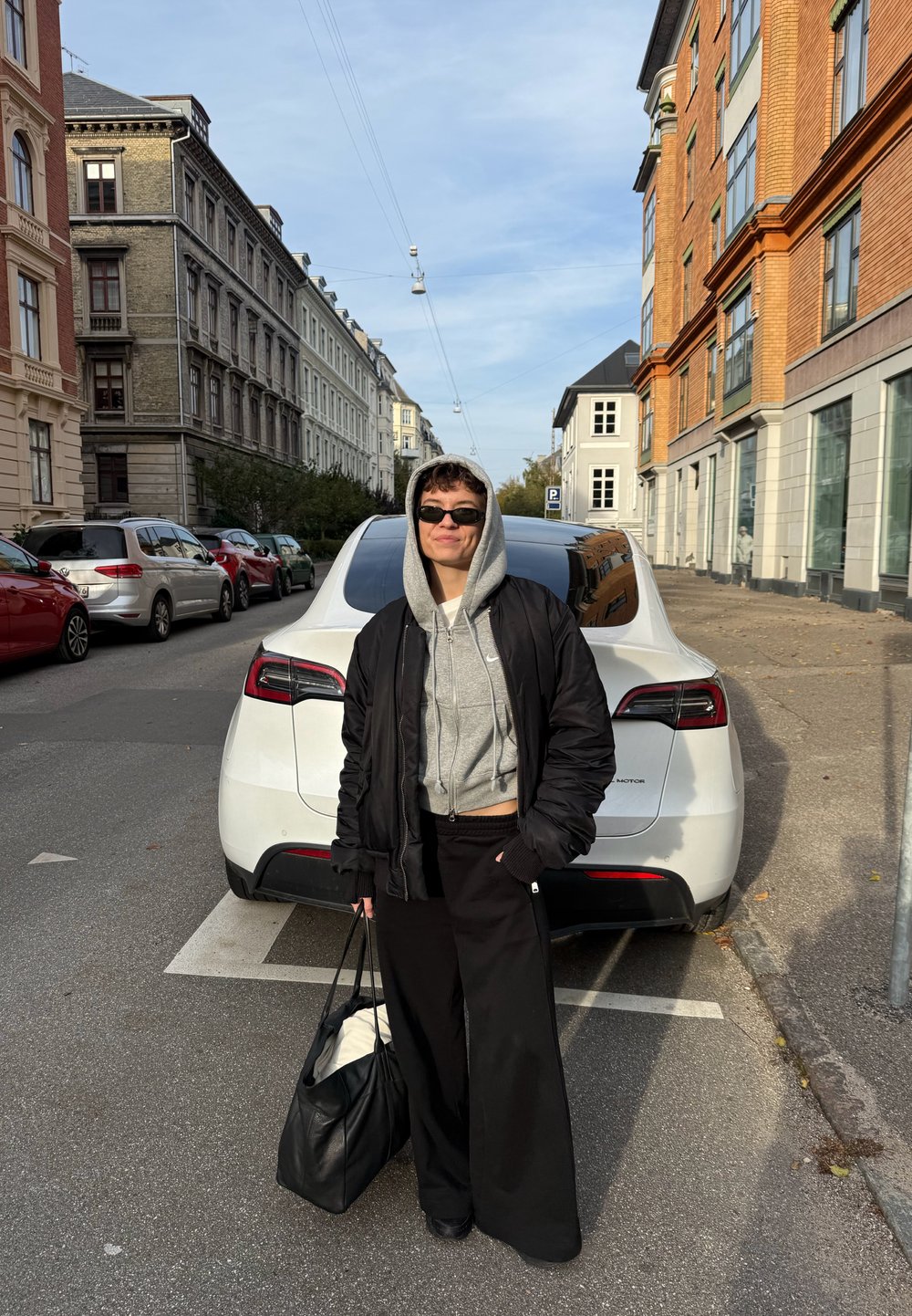 Inimene, kes kannab musta jope, halli kapuutsi ja prille, seisab tänaval musta koti käes, valge auto ees, millel on parkimise koht linna alal.