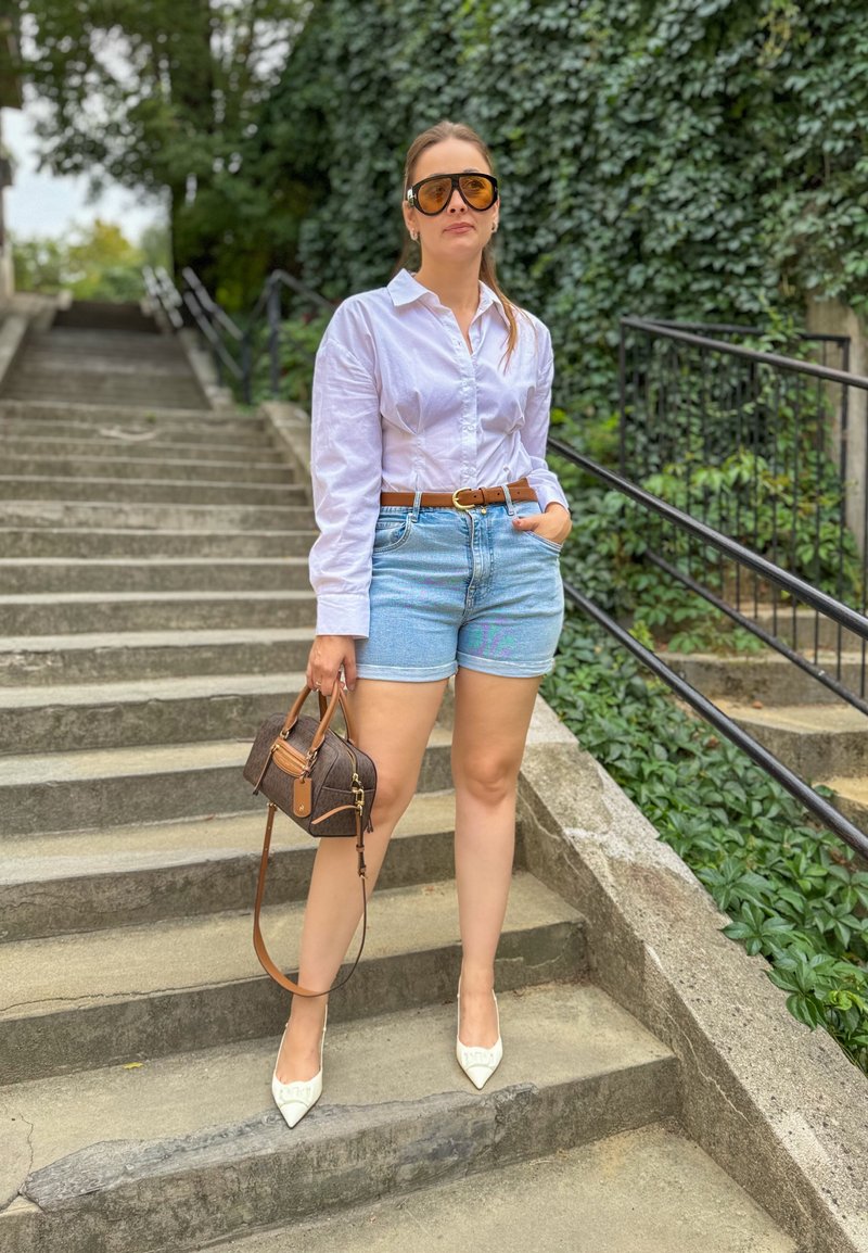 Femme portant des lunettes de soleil surdimensionnées, une chemise blanche, un short en denim et des talons blancs, debout sur des escaliers extérieurs en béton avec un fond végétal.