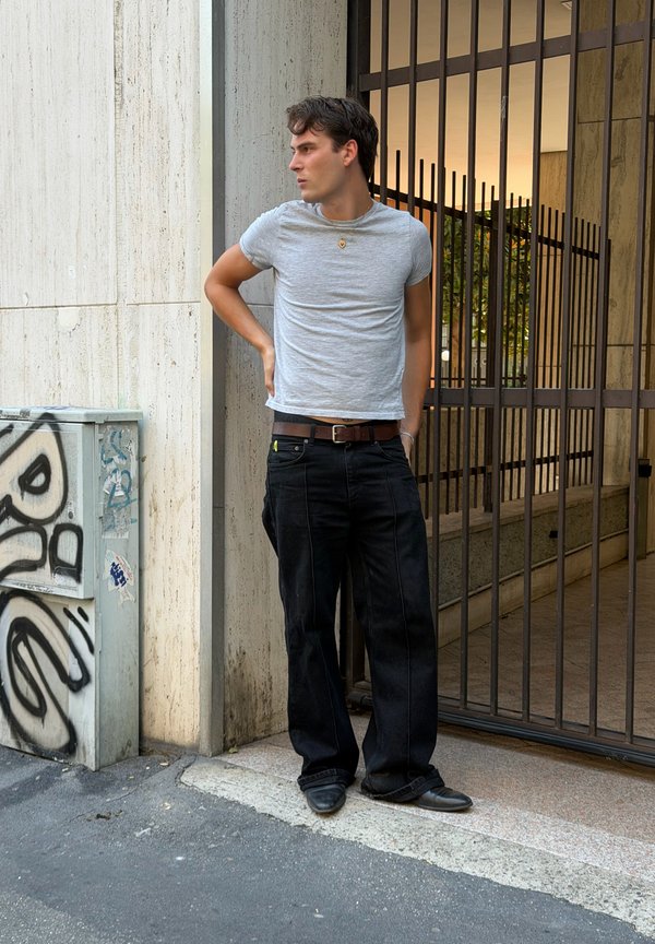Jeune homme en t-shirt gris et pantalon noir debout avec une main dans le dos près d'une entrée grillagée et d'un boîtier utilitaire couvert de graffitis.