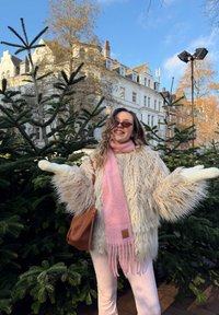 Textured cream faux fur coat with wide sleeves, paired with a pink scarf, light-coloured trousers, and a brown handbag, surrounded by evergreen trees.