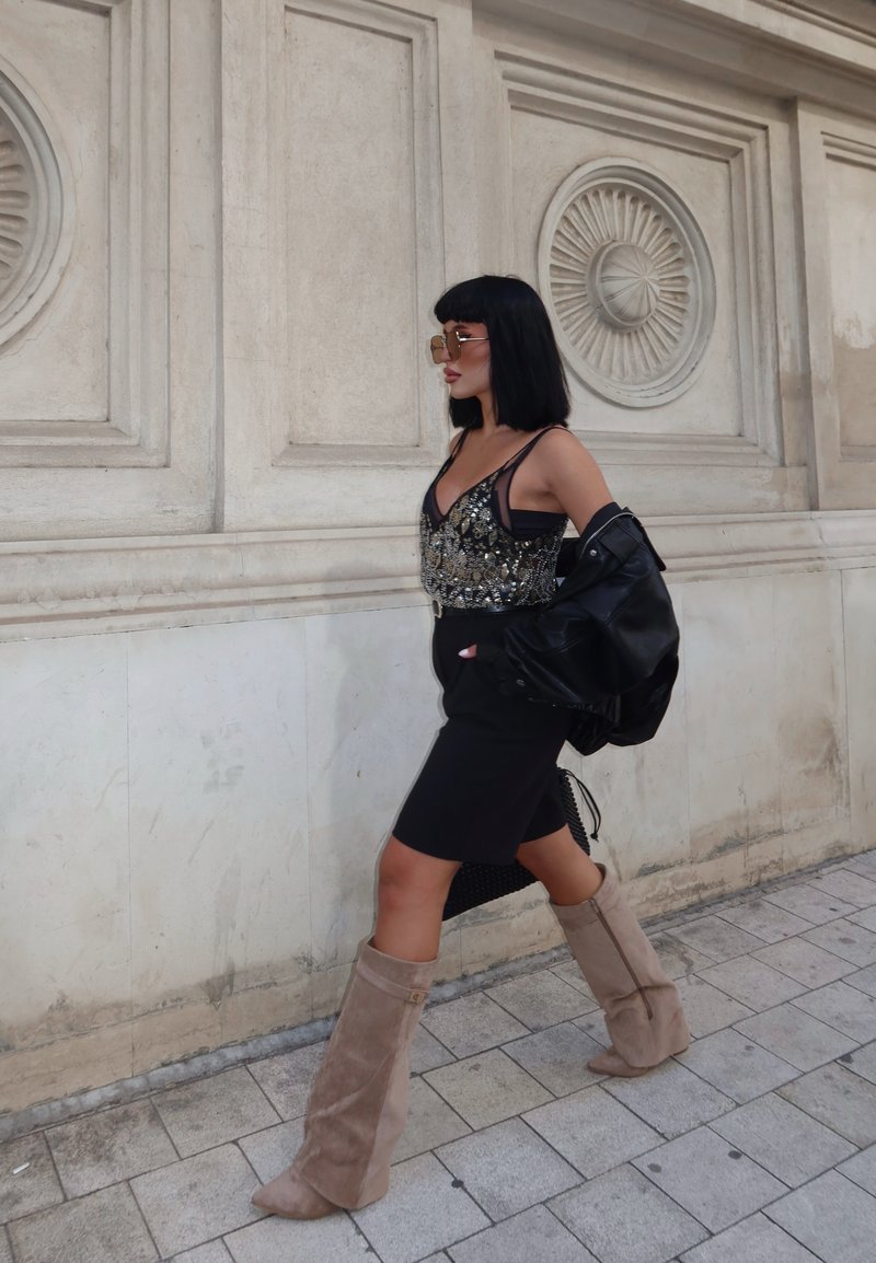 Robe noire avec un corsage orné de bijoux, associée à un blouson en cuir noir. Bottes beiges jusqu'aux genoux, grandes lunettes de soleil. En train de marcher près d'un mur orné.