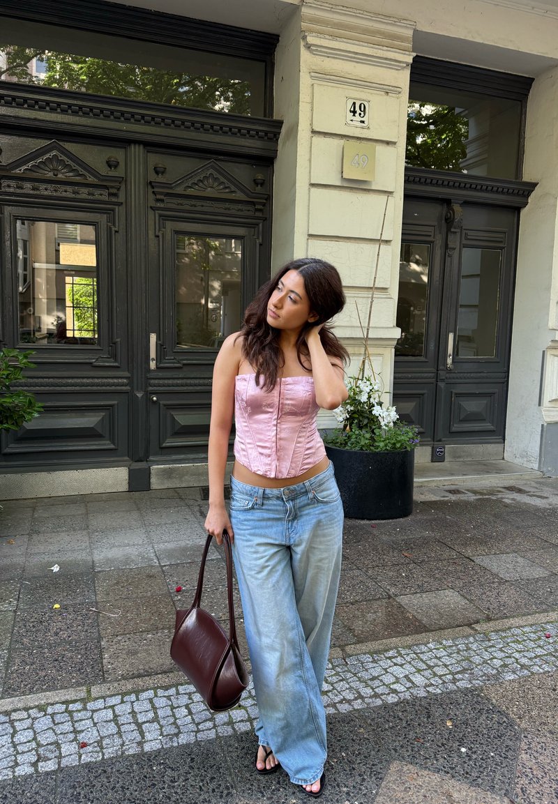 Jeune femme en corset rose et jeans larges tenant un sac à main marron se tient sur le trottoir de la ville près de portes en bois foncé et de plantes en pot.