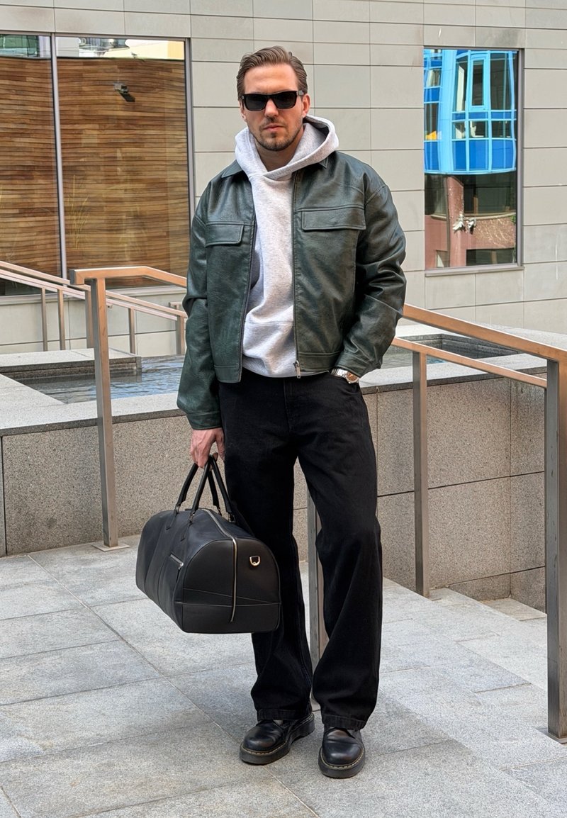 Homme portant des lunettes de soleil, un sweat à capuche gris, une veste en cuir verte, un pantalon noir et des chaussures, tenant un sac de sport noir, debout près des rampes métalliques à l'extérieur.