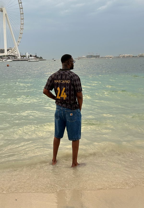Sort og guld ternet skjorte med korte ærmer med "MARCANO 24" trykt på ryggen, parret med blå denimshorts, stående på våd sand.