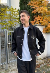 Must musta ülikoolitüki jope valgete triipudega mansettidel, trukkidega ja tikitud logoga. Kandatud valge särgi kohal, taustal sügislehtede värvid.