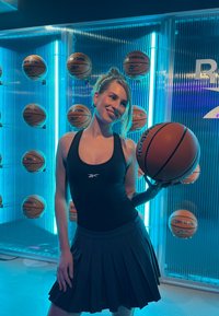 Γυναίκα με μαύρη αθλητική φόρμα κρατάει μια μπάλα μπάσκετ μπροστά από έναν τοίχο με πολλές μπάλες μπάσκετ και μπλε νέον φωτισμό.