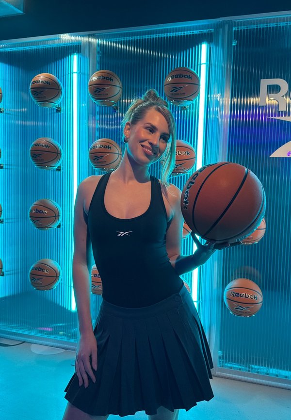 Femeie îmbrăcată în echipament sportiv negru, ținând un baschet în fața unui perete cu mai multe mingi de baschet și iluminare neon albastră.