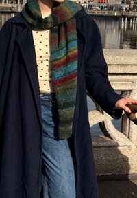 Garš svītrains šalle ar krāsām, tostarp zaļu, zilu un sarkanu, valkāta virs tumši zilas jakas un polka punktu krekla, kopā ar gaiša džinsa džinsiem.