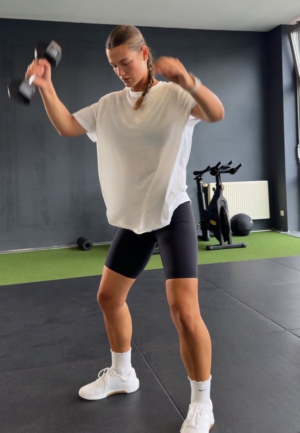 Femme en chemise blanche et short noir soulevant des haltères dans une salle de sport avec un vélo d'appartement et un ballon médicinal en arrière-plan.