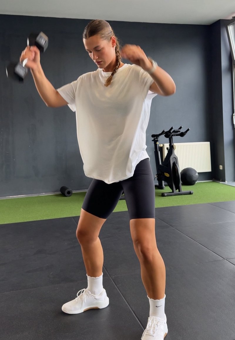 Femme en chemise blanche et short noir soulevant des haltères dans une salle de sport avec un vélo d'appartement et un ballon médicinal en arrière-plan.